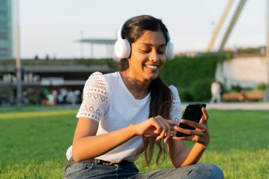 Beautiful smiling Indian woman holding mobile phone listening music in wireless headphones. Happy asian female hipster using mobile app shopping online, making reservation. Mobile banking concept