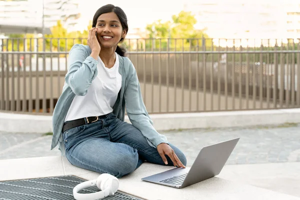 Cheerful smiling Indian woman using laptop computer, talking on mobile phone working project sitting at workplace
