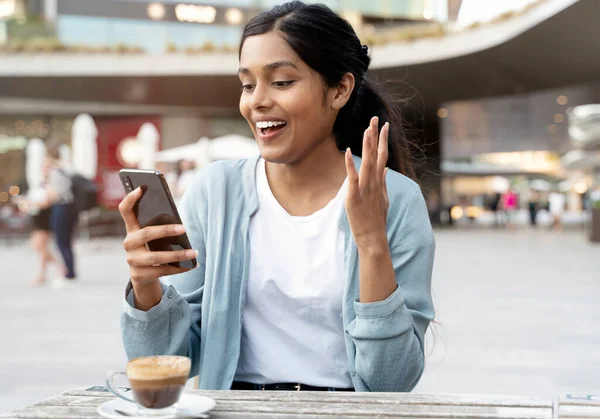 Young excited Indian woman using mobile phone shopping online with sale sitting in cafe. Happy hipster female playing mobile game. Asian student reading exam results