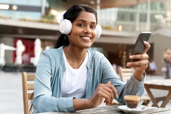 Smiling Indian woman wearing wireless headphone using mobile phone listening music looking at camera. Young confident asian female communication online drinking coffee sitting in cafe