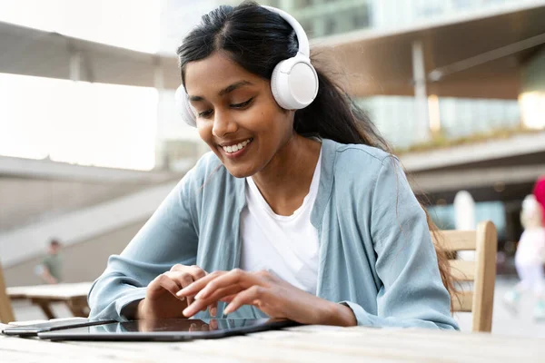Dijital tablet kullanan, kablosuz kulaklık takan, video izleyen, modern kafede iletişim kuran gülümseyen Hintli kadın. Sosyal medya kavramı  