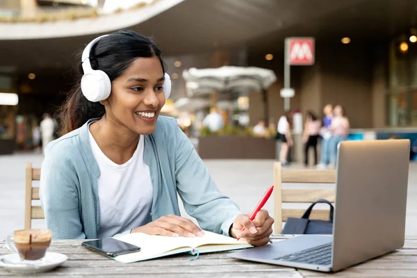 Smiling Indian woman using laptop computer studying, taking notes, exam preparation, online education concept. Young asian freelancer working project, watching training courses sitting at workplace