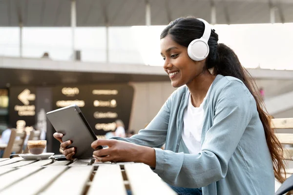 Smiling Indian woman using digital tablet wearing wireless headphones watching training courses, communication online in modern cafe. Happy asian student studying, distance learning, online education