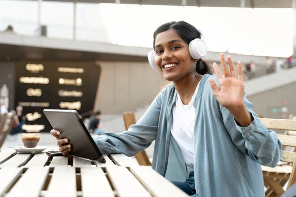 Smiling Indian student waving hand using digital tablet, studying, distance learning, online education concept. Portrait of happy successful asian freelancer wearing headphones looking at camera