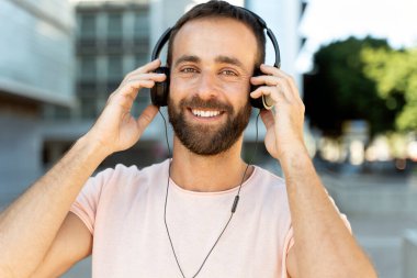 Yakışıklı, sakallı, kulaklık takan, müzik dinleyen, caddede kameraya bakan adam. 