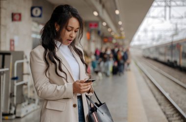 Tren istasyonunda cep telefonu kullanan güzel, melez bir iş kadını, kopyalama alanı. Akıllı telefon ekranına bakan düşünceli turist treni bekliyor. Ulaşım, seyahat kavramı 