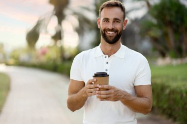 Handsome smiling hispanic man wearing white t shirt holding cup of coffee looking at camera standing in park, copy space. Coffee break concept 