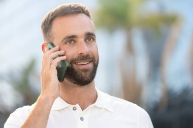 Close up portrait of handsome smiling latin man talking on mobile phone on the street, looking away Copy space 