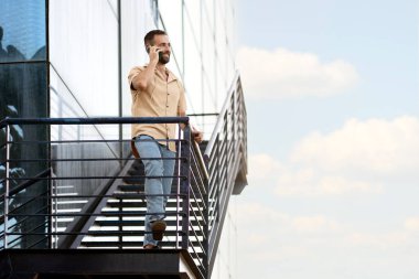 Confident smiling hispanic businessman talking on mobile phone  standing in office against blue sky. Copy space. Career, successful business concept 