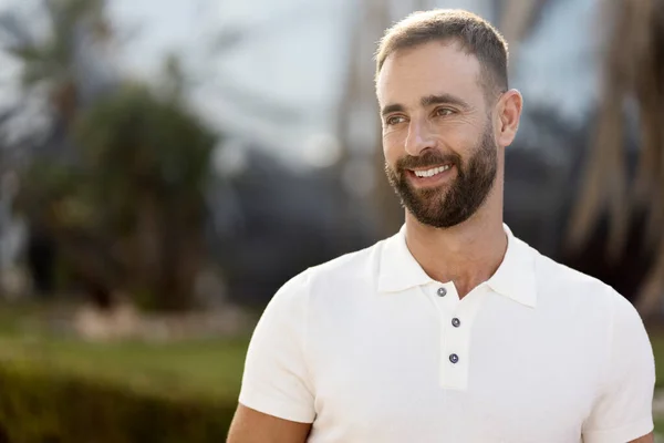 Confident smiling hispanic man wearing white stylish t shirt looking away standing on the street, copy space. Portrait of middle aged bearded fashion model posing for pictures, sunny day