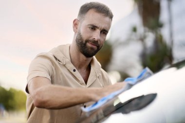 Yakışıklı düşünceli İspanyol adam mikro fiber bez kullanarak arabasını temizliyor.