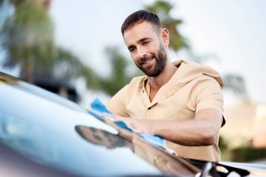 Yakışıklı gülümseyen adam mikro fiber bez kullanarak arabasını temizliyor. Araba yıkama servisi konsepti