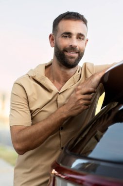 Gülümseyen sakallı latin adam yeni arabanın yanında durmuş kameraya bakıyor.