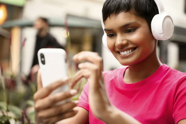 Smiling asian woman wearing wireless headphones listening music on the street. Indian hipster female using mobile phone shopping online, text messaging, playing mobile game outdoors