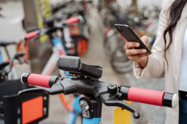 Mobil uygulama kullanan kadın elinin kapatılması, sokakta kiralık elektrikli scooter için QR kodu taraması, seçici odaklanma. Eko taşımacılığı