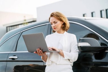 Kafkasyalı ciddi işadamı dizüstü bilgisayarını kullanıyor ve arabanın yanında duruyor.  