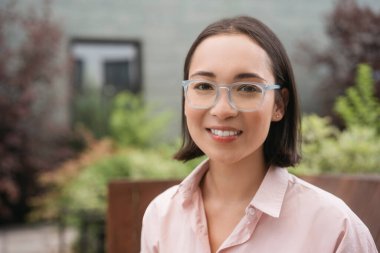 Üniversite kampüsünde kameraya bakan şık gözlüklü, zeki, gülümseyen Asyalı öğrenci portresi, eğitim konsepti.