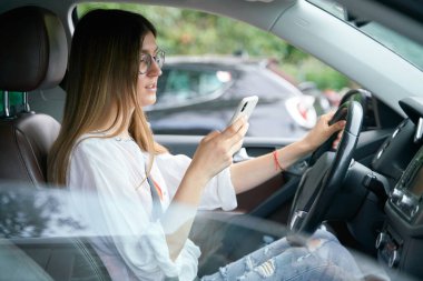 Gözlüklü çekici bir kadın arabada oturuyor ve akıllı telefon ekranına bakıyor. İş kadını araba kullanmakla meşgul 