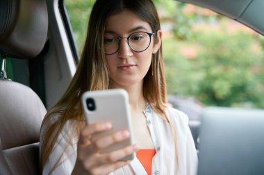 Beyaz kadın lüks bir arabada oturmuş, akıllı telefon ekranına bakıyor, internetten iletişim kuruyor. Ulaşım kavramı