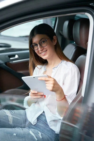 Vertical shot of the beautiful woman using digital tablet sitting in car, working online. Technology concept