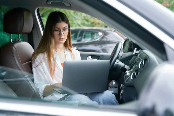 Successful confident businesswoman sitting in the driver seat of the car using laptop computer, typing, working online. Beautiful female in car, transportation concept