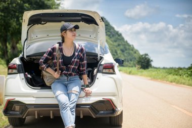 A female traveler with a backpack in a hatchback on a road with mountains, trees and sky in the background.