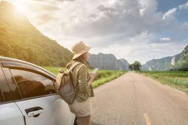 A young woman backpacking beside a car and using a smartphone on a tourist road with mountains and sky in the background.