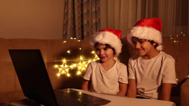 Karácsonyi üdvözlet online. Két kisfiú a Télapóban egy laptopot kalapol. Videóhívások fogadása barátoknak, rokonoknak és szülőknek. A karácsony fogalma — Stock videók