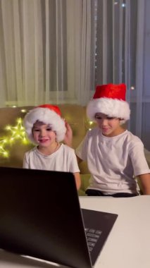 Noel selamları çevrimiçi. Noel Baba 'daki iki küçük çocuk bir dizüstü bilgisayara şapka takıyor. Arkadaşlara, akrabalara ve ailelere video görüşmeleri. Noel konsepti. Dikey video