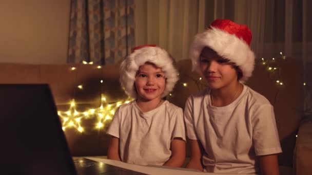 Noel selamları çevrimiçi. Noel Baba 'daki iki küçük çocuk bir dizüstü bilgisayara şapka takıyor. Arkadaşlara, akrabalara ve ailelere video görüşmeleri. Noel konsepti — Stok video