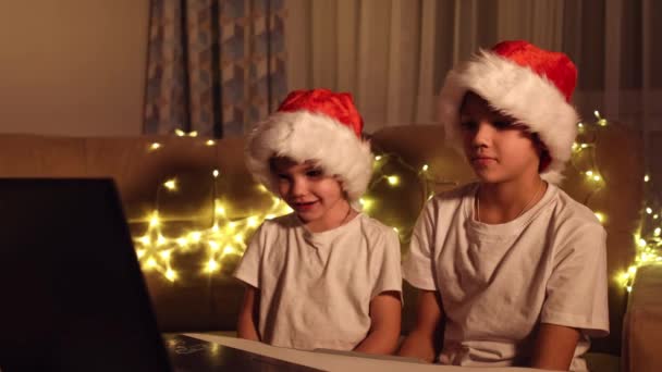 Saludos de Navidad en línea. Dos niños pequeños en Santa Claus sombreros de un ordenador portátil. Saludo a las videollamadas a amigos, familiares y padres. El concepto de Navidad — Vídeo de stock