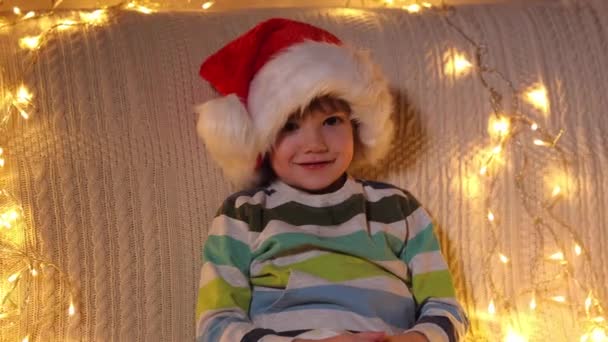 Un petit garçon dans un chapeau de Père Noël regarde la caméra et sourit à l'arrière-plan des lumières de vacances. Noël. Au ralenti. Émotions positives des gens. — Video