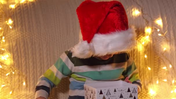 Un garçon ouvrant boîte cadeau à l'arrière-plan des lumières de vacances. Noël. Au ralenti. Émotions positives des gens. — Video
