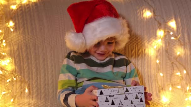 Un garçon ouvrant boîte cadeau à l'arrière-plan des lumières de vacances. Noël. Au ralenti. Émotions positives des gens. — Video