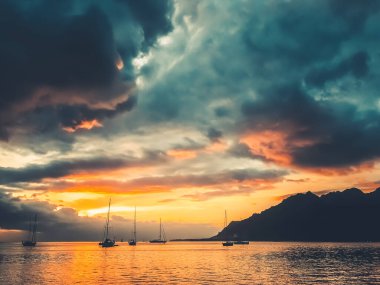 Dramatic nature sunset in tropical island lagoon. Yachts silhouette against bright orange blue clouds in sky. Ocean calm water and mountain hill. Wonderful picturesque scene. Majestic travel landscape