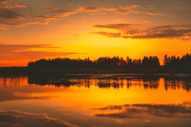 Bright orange sunset over lake, golden clouds and silhouette forest reflect in the water. Amazing nature background texture