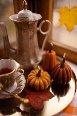 Balkabağı şeklinde mumlar, çay fincanı, penceresinde sonbahar yaprakları, çaydanlık, altın tepsi ve klasik kitapla sonbahar durgun hayatı.