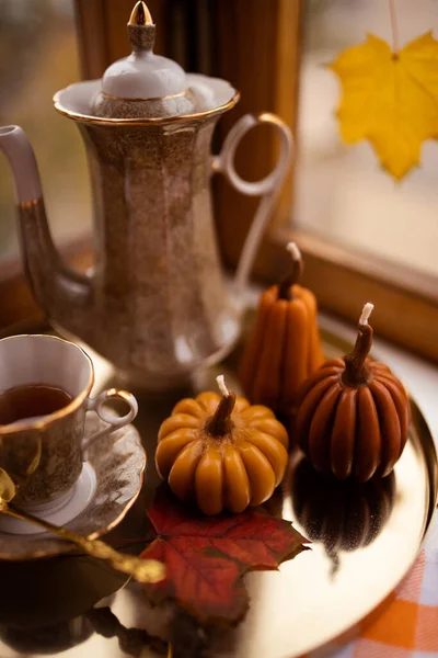 Balkabağı şeklinde mumlar, çay fincanı, penceresinde sonbahar yaprakları, çaydanlık, altın tepsi ve klasik kitapla sonbahar durgun hayatı.