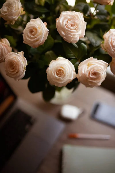 Workplace with a dark grey laptop, beige roses bouquet, a green notebook, a mobile phone, an orange pan on the wooden table