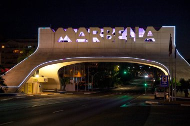 Marbella, İspanya - 21 Temmuz 2020: Marbella işareti, şehrin girişinde, gece perspektifi. Işıklar açık, tabelayı farklı renklerle aydınlatıyor. Uzun pozlu fotoğrafçılık. araba ışıkları  