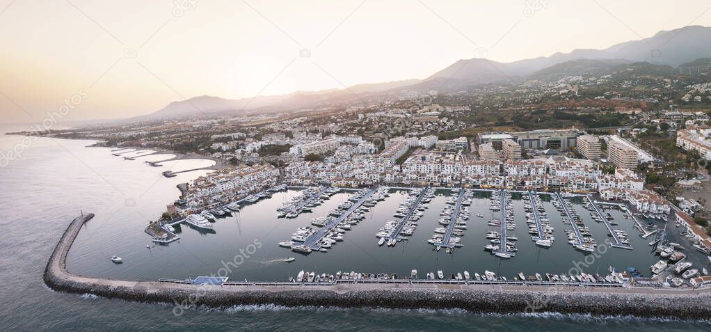 Aerial panorama of Puerto Banus Bay - Marbella - Spain. Beautiful golden hour colours. View of sun going down in mountain from Sierra Blanca. Luxury and famous travel destination. Expensive yachts