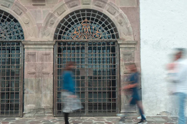 İnsanlar gün boyunca metal bir çitle, kemerli taş bir kapının önünde hızla yürüyorlar.
