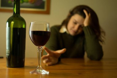 Close-up on depressed woman with green jacket sitting alone at a table trying to reach glass of red wine with her hand