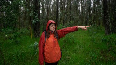 Siyah sırt çantalı ve kırmızı su geçirmez ceketli İspanyol bir kadının gün ortasında bir ormanın ortasında uzanan koluyla yönünü göstermesi.