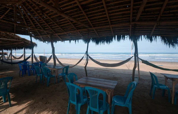 View of a rustic restaurant without people, with a bamboo roof, wooden tables, plastic chairs and hammocks hanging from poles by the beach on a sunny day