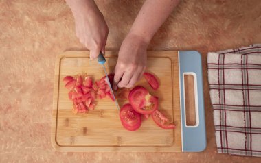 Top view of woman's hands chopping tomato slices with a blue knife on a wooden board at the kitchen counter
