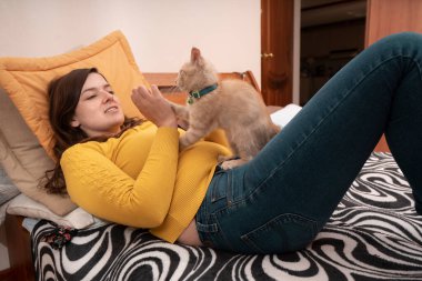 Beautiful Hispanic young woman smiling dressed in yellow jacket, lying on her bed playing with her little kitten with her hands