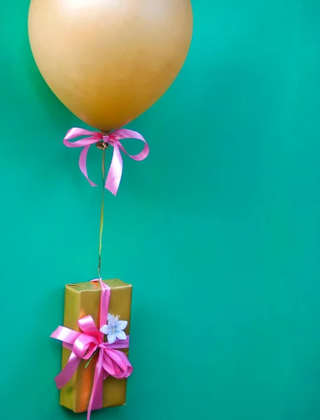 A golden balloon with a pink ribbon and a gift in a golden package with a pink ribbon and a white flower in the middle on a turquoise background