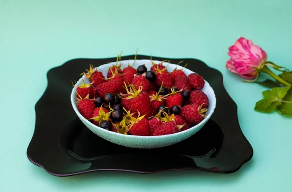 Raspberries and black currants in a bowl on a black plate with a rose in the background