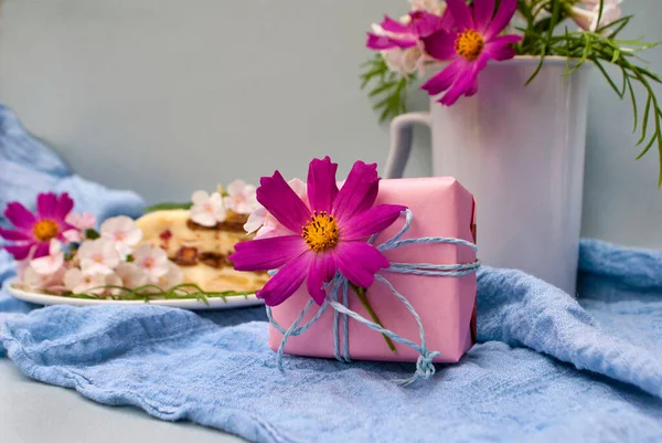 A small light pink gift with a flower and a piece of cake surrounded by flowers on a blue fabric